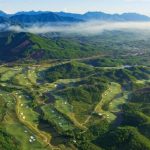 Aerial view of golf fairways and bunkers among green hills beneath a misty mountain ridge