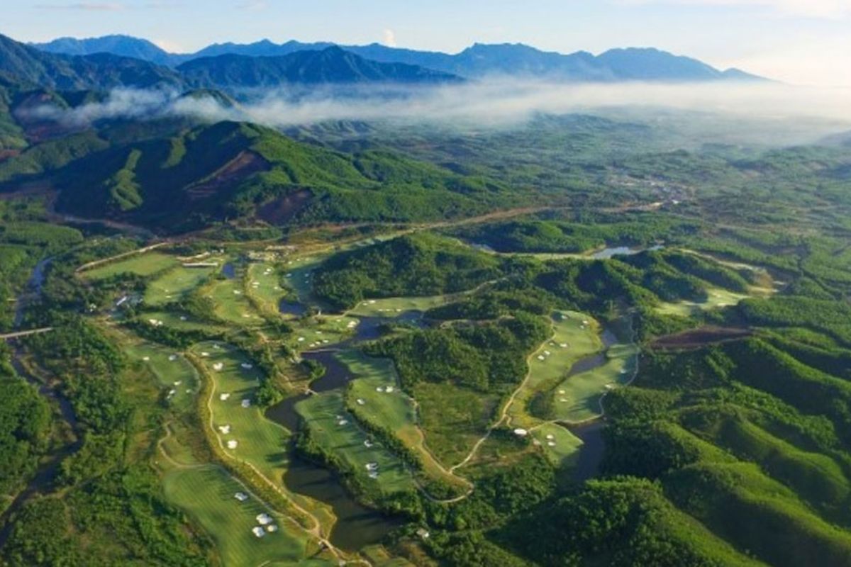 Aerial view of golf fairways and bunkers among green hills beneath a misty mountain ridge